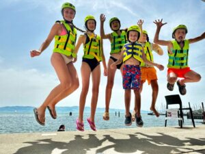 Kinder in Badekleidung,Schwimmwesten und Helmen hüpfen in die Luft, im Hintergrund der Bodensee