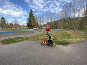 Kind mit Helm fährt Fahrrad auf einem Spielplatz im Allgäu, Natur im Hintergrund.